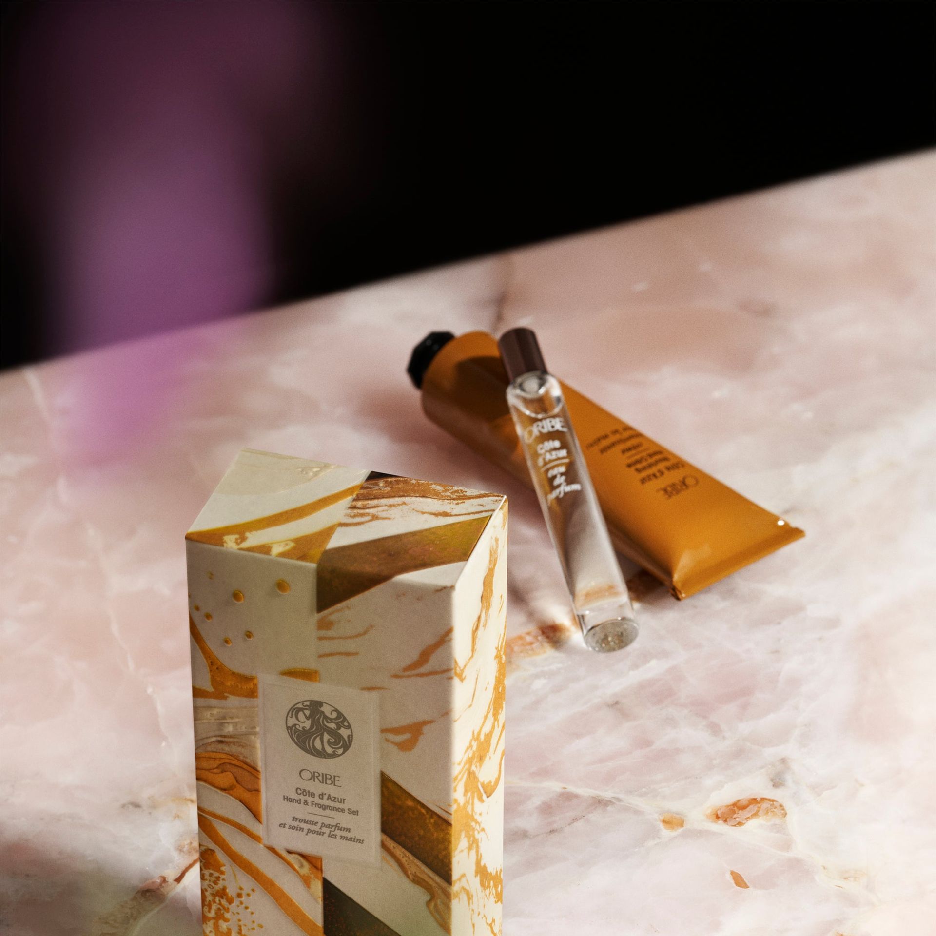 A branded Oribe product box, a gold tube, and a small glass vial rest on a polished pink marble surface.