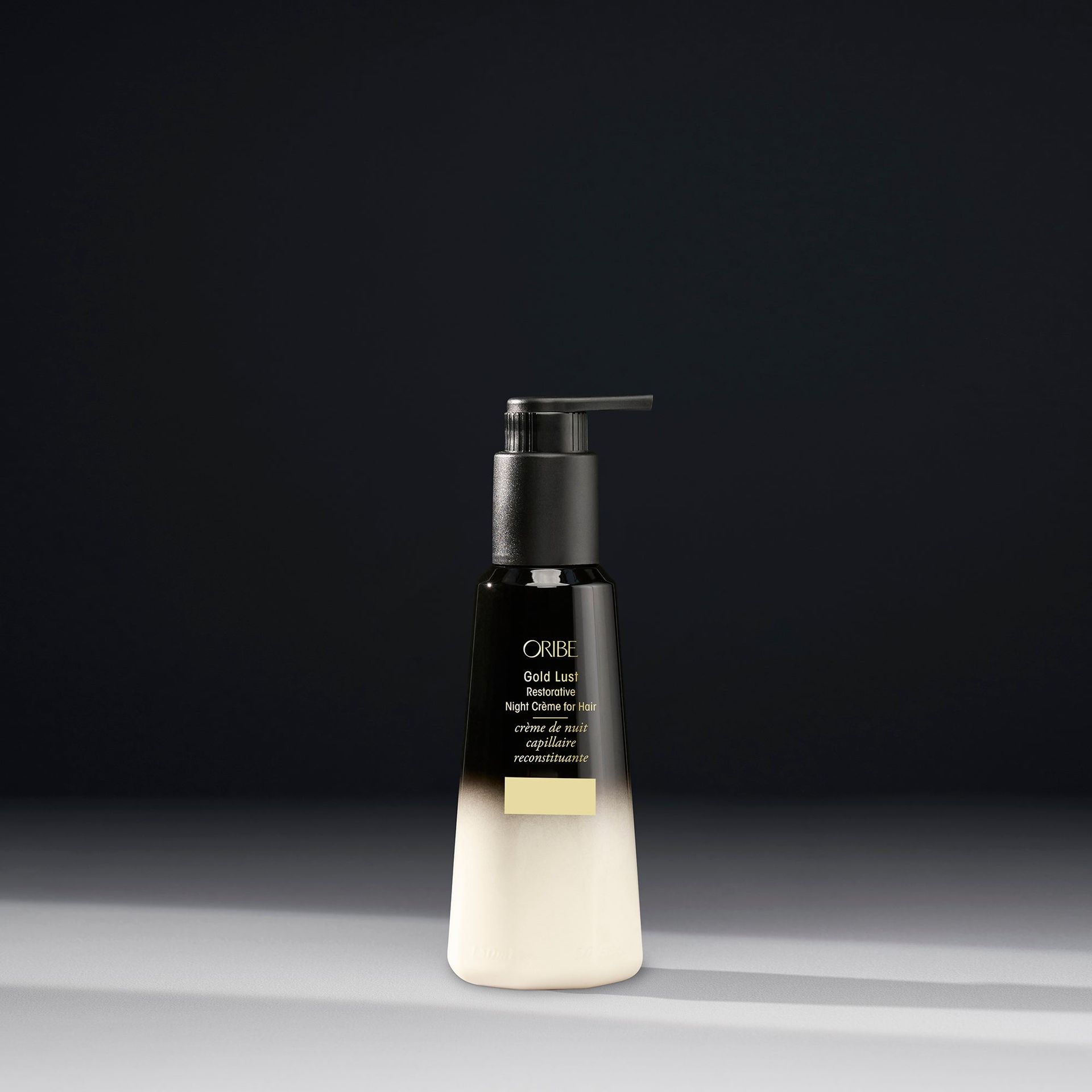 A black and white ombre pump bottle of Oribe Split End Seal hair treatment stands on a grey surface against a dark wall.