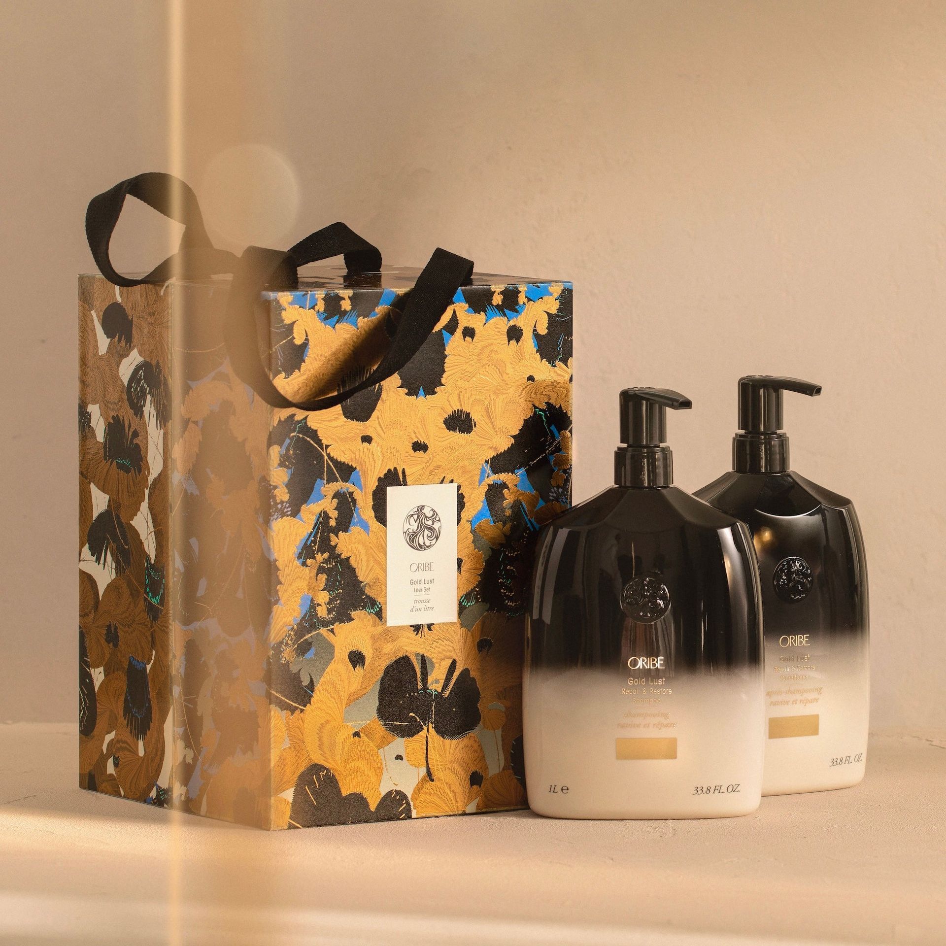 A gift bag with a black and gold floral pattern next to two pump-dispenser bottles with a black-to-white gradient finish.
