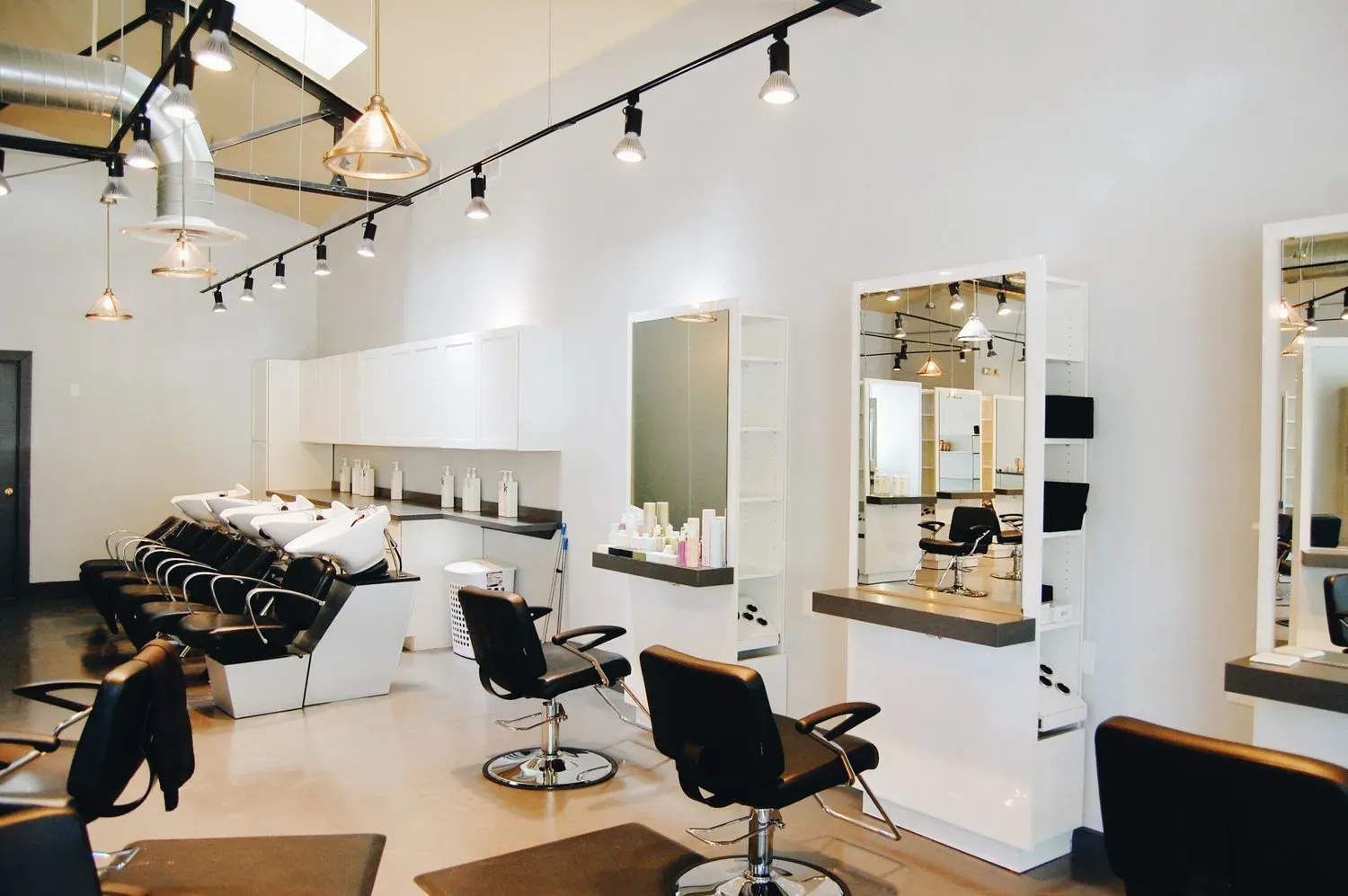 An empty hair salon with black styling chairs, washing stations, white counters, and overhead track lighting.