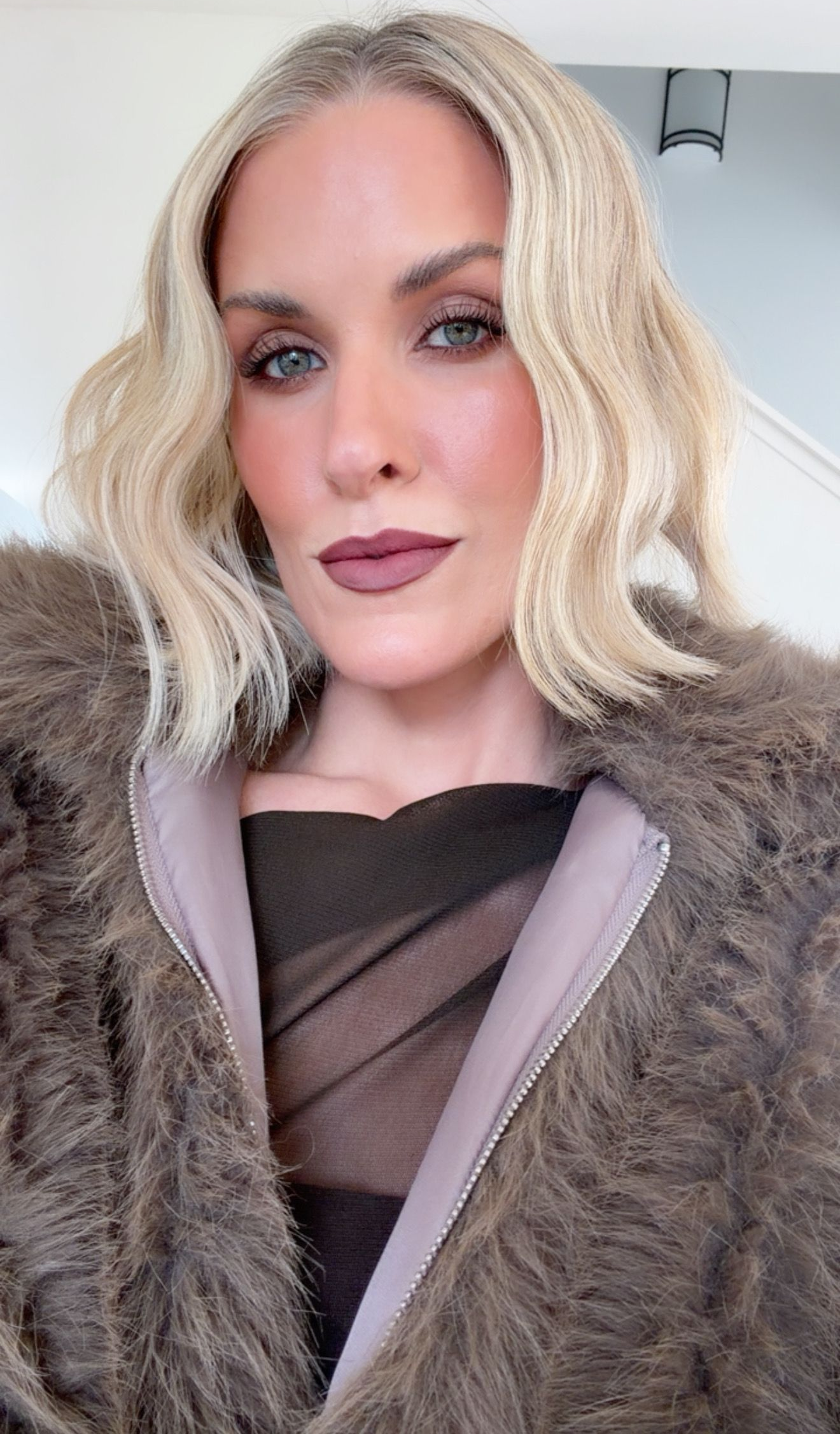 A person with wavy blonde hair wears a dark, fuzzy coat over a brown top, posing for a portrait with a neutral expression.