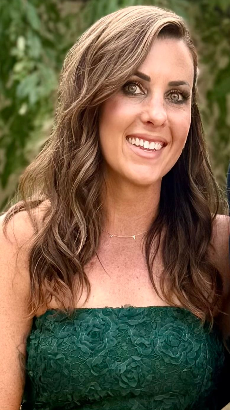 A smiling person with long brown hair wearing a textured, green strapless top against a blurred outdoor background.
