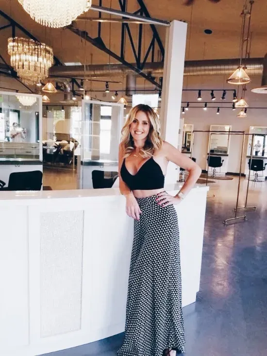 A person smiles, leaning on a white counter in a modern, well-lit hair salon with pendant lights and floor-length skirts.