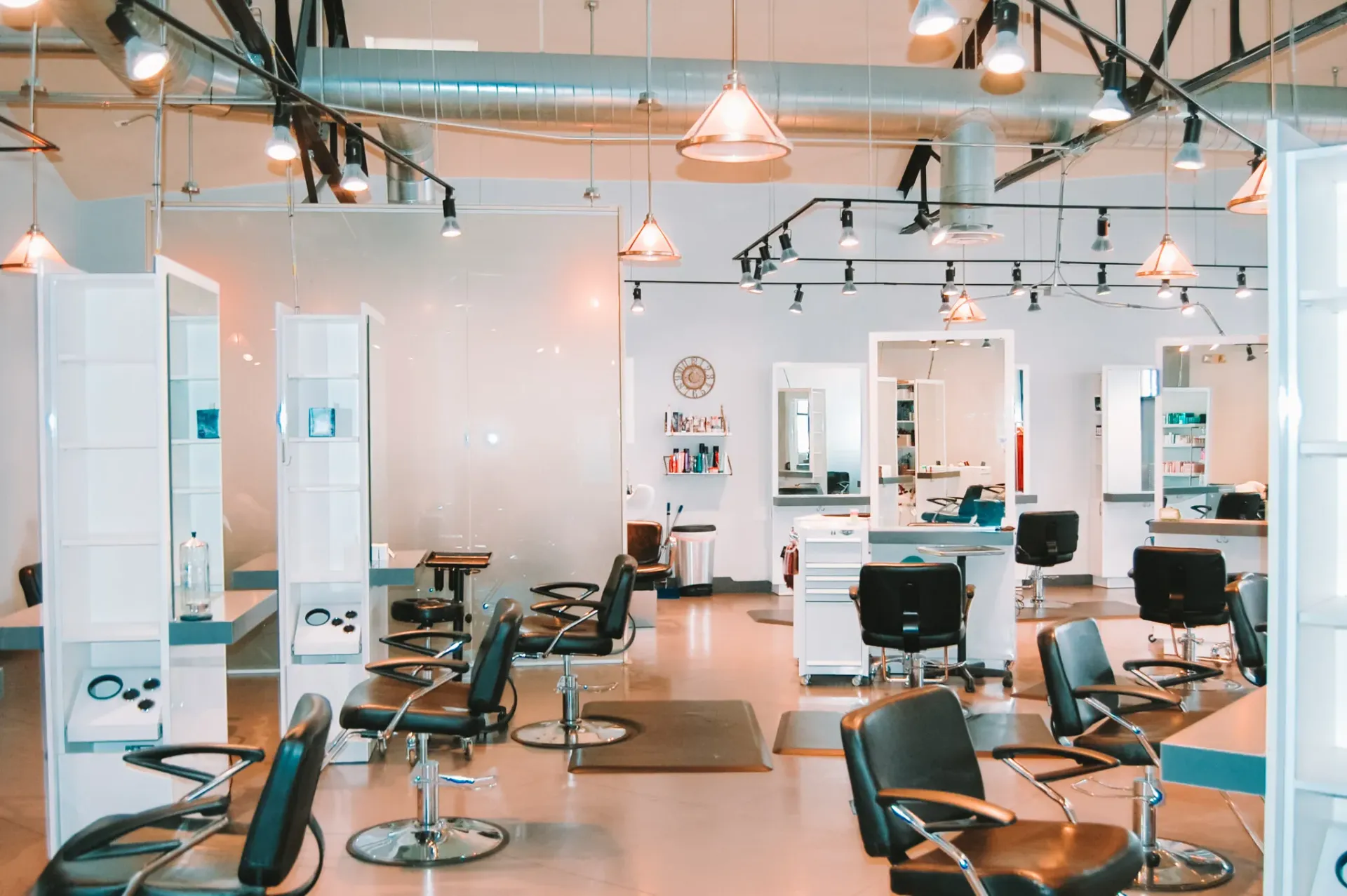 A bright, modern hair salon with multiple styling stations, black chairs, large mirrors, and hanging pendant lights.