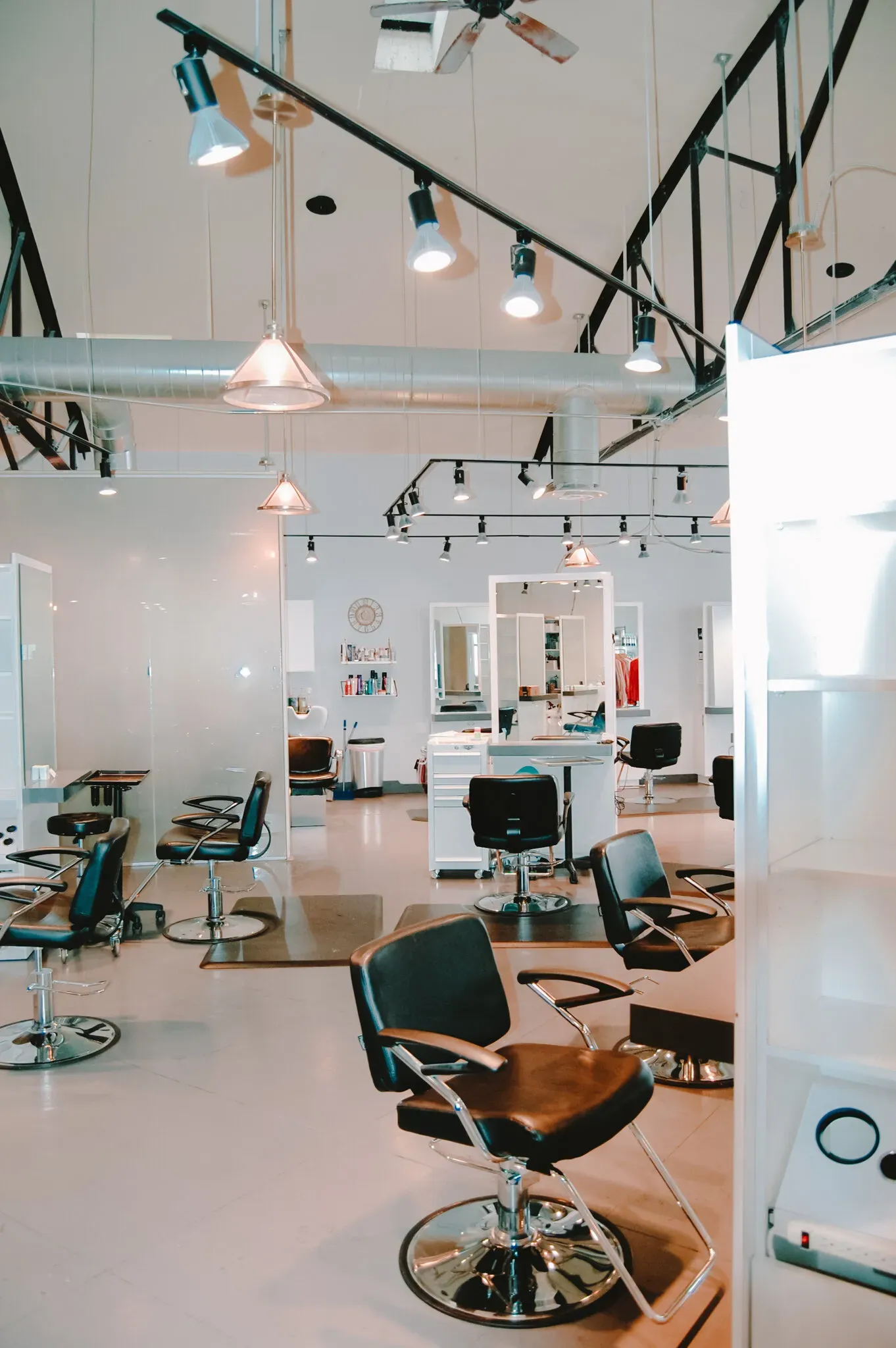 A bright, modern hair salon with multiple styling stations featuring black chairs, mirrors, and industrial track lighting.