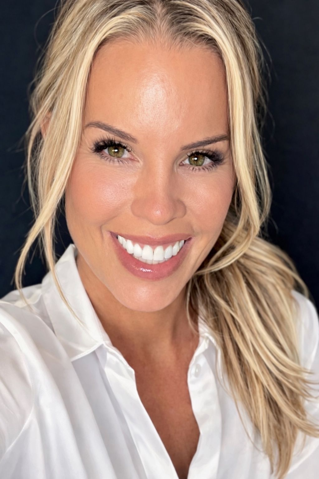 A person with blonde hair pulled back, smiling and wearing a white collared shirt against a dark background.