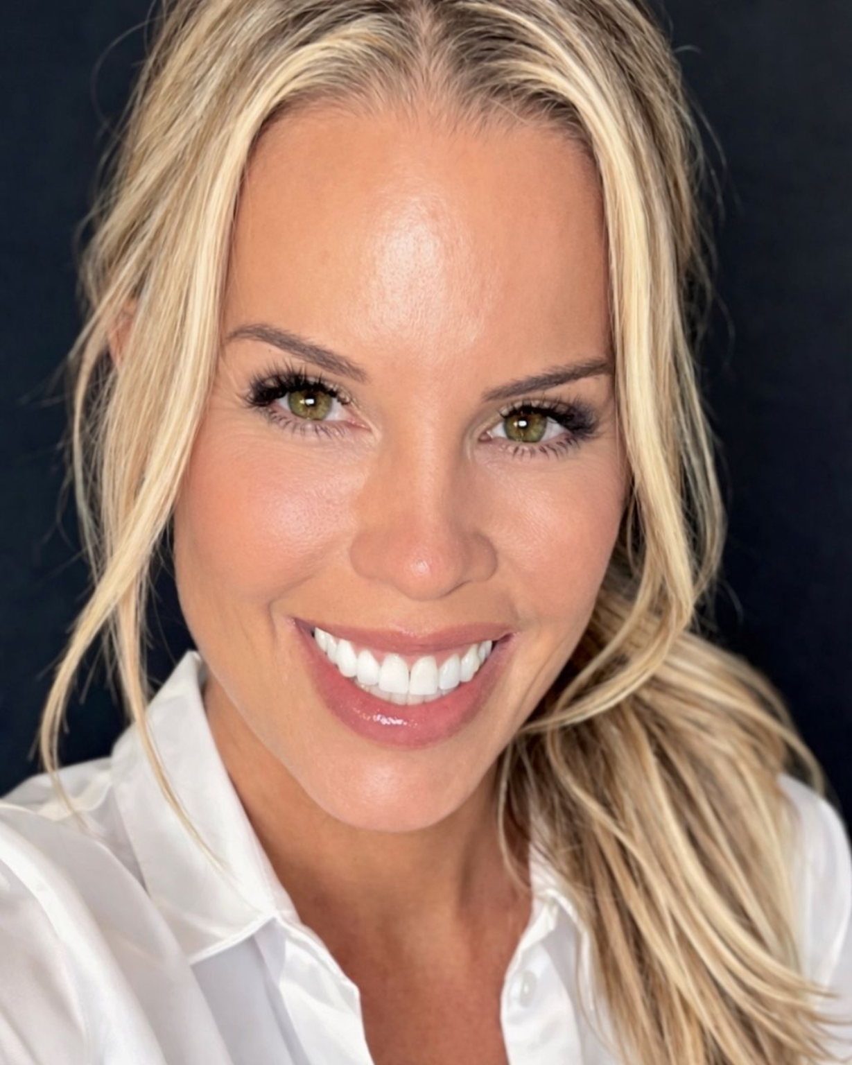 A person with blonde hair in a ponytail, wearing a white collared shirt against a black background, smiling at the camera.