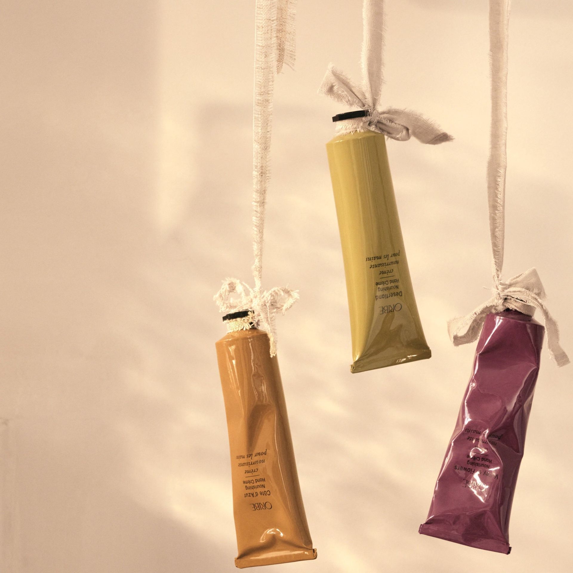 Three colorful tubes—mustard, gold, and purple—hang by ribbons against a light, neutral background.