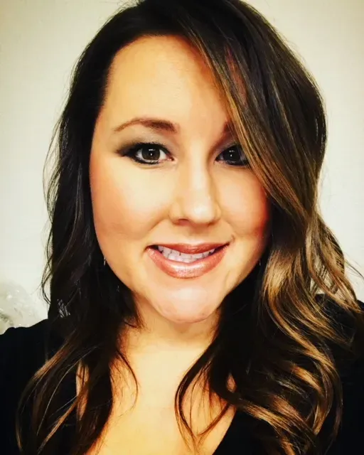 A headshot of a person with long, dark, wavy hair with lighter highlights, smiling at the camera against a white background.
