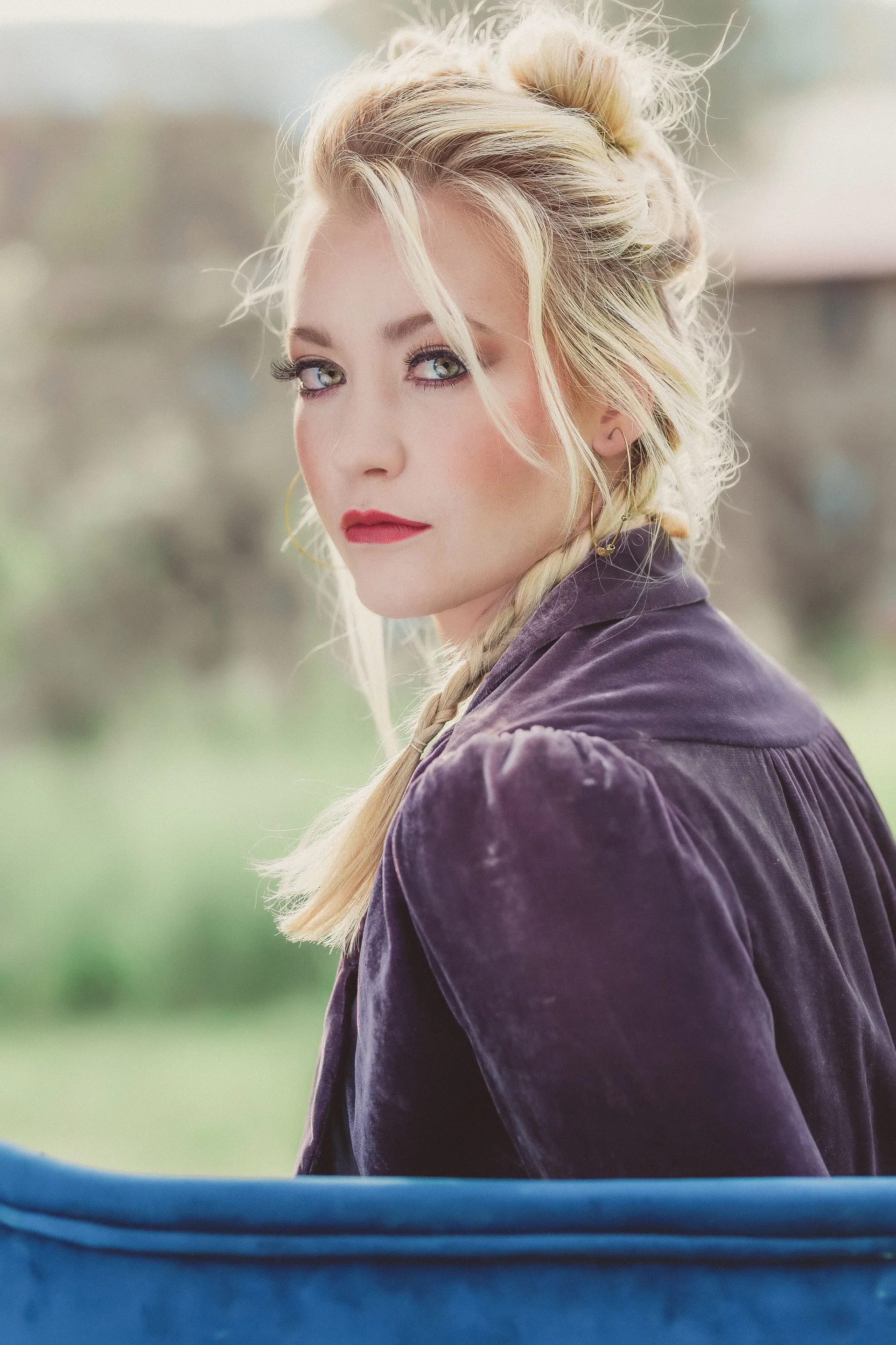 A blonde individual with braided hair looking over their shoulder, wearing a dark purple top against a blurred background.