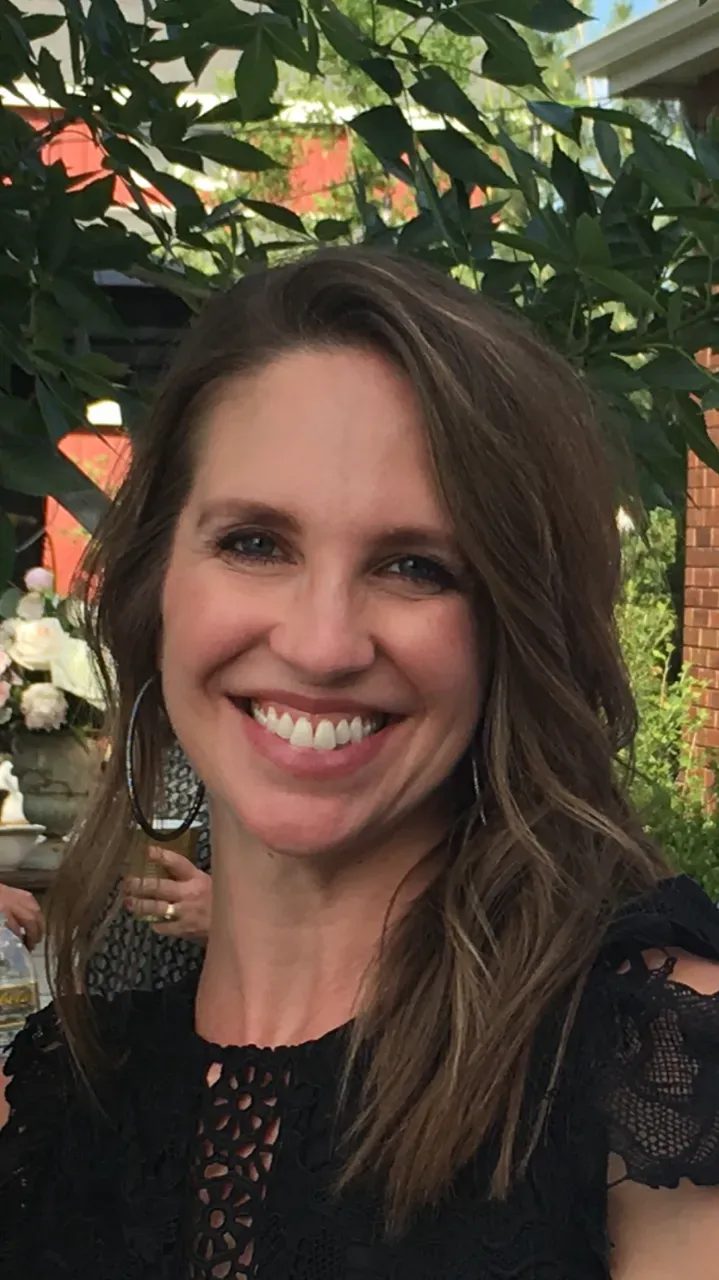 A smiling woman with shoulder-length wavy brown hair wearing a black top, positioned in an outdoor garden setting.
