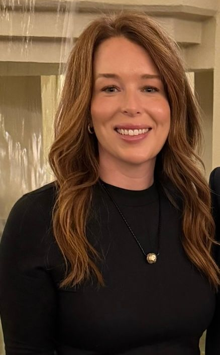 A person with wavy brown hair smiling at the camera, wearing a dark top, with a blurred outdoor background.