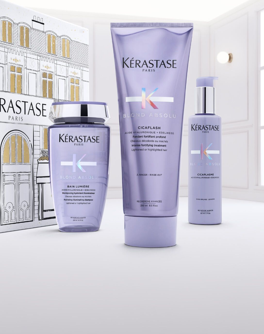 Three Kérastase hair care products in lavender packaging stand before a matching gift box against a white background.
