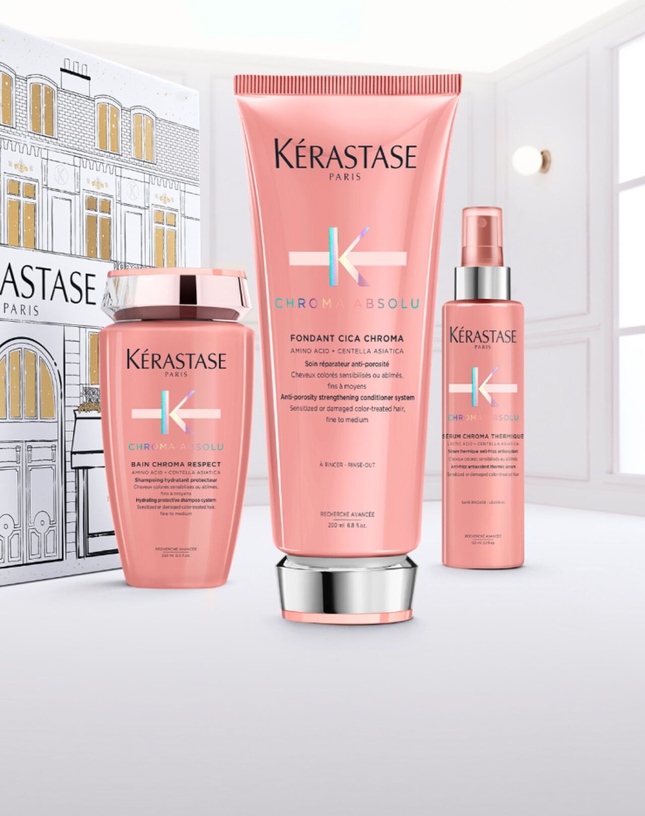 Three pink Kérastase hair care bottles arranged on a white surface against a matching branded box and a bright room.