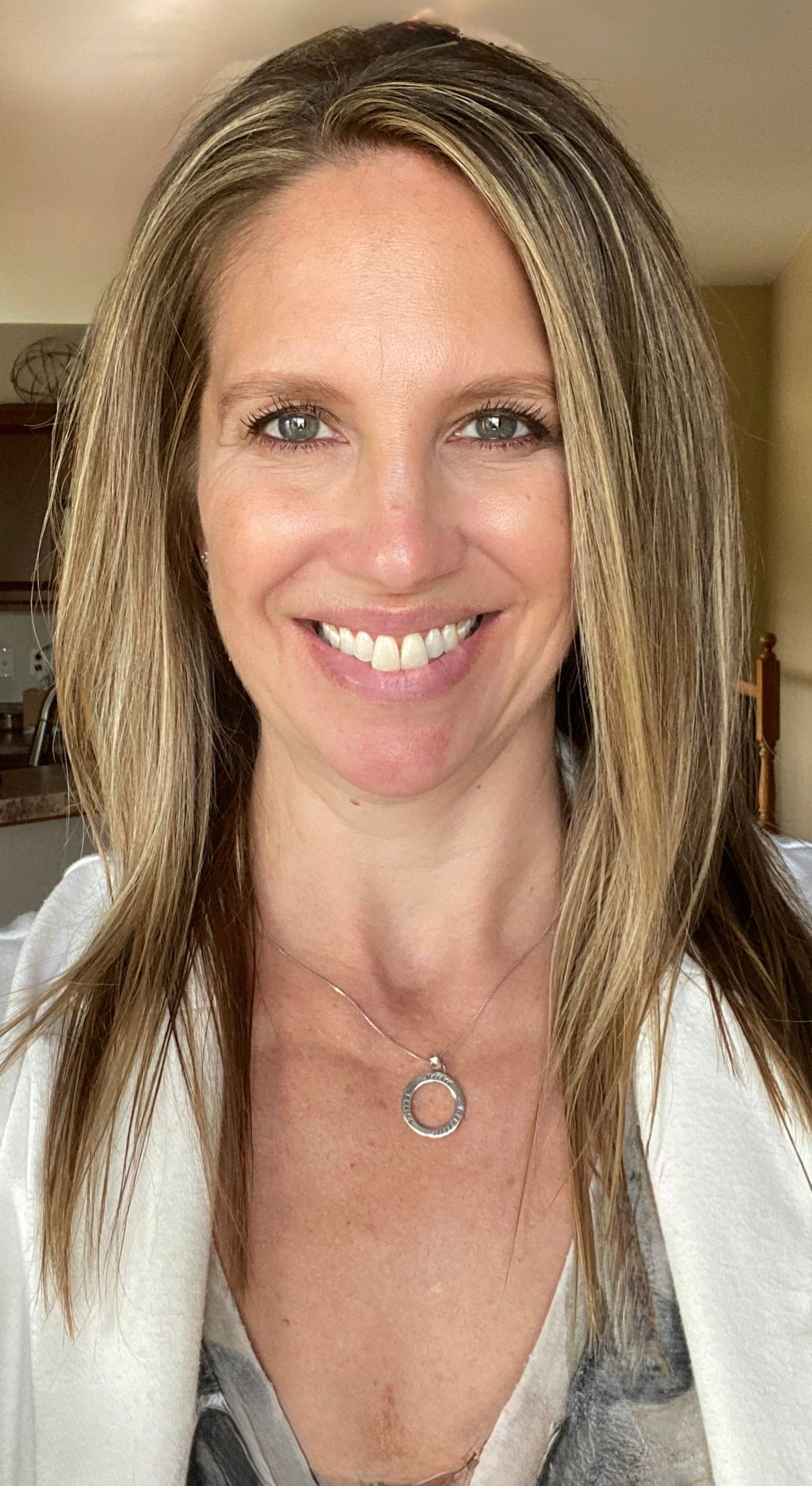 A smiling person with shoulder-length, textured hair, wearing a silver pendant necklace and a white jacket.