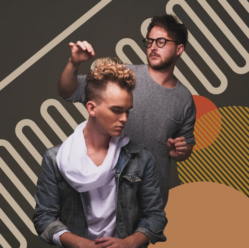 A stylist with glasses shapes the hair of a person wearing a denim jacket and white scarf against a patterned background.