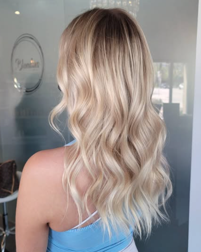 Back view of long, wavy blonde hair with dark roots, styled in soft curls against a bright indoor background.
