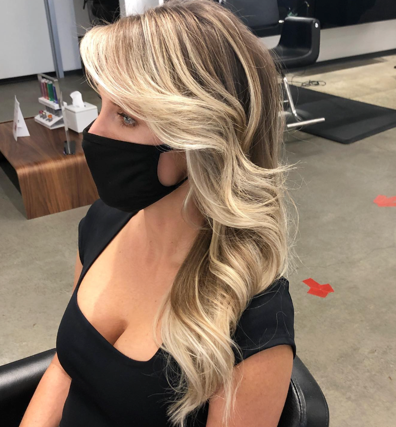 A person with long, wavy blonde hair styled in a salon, wearing a black top and a black face mask.