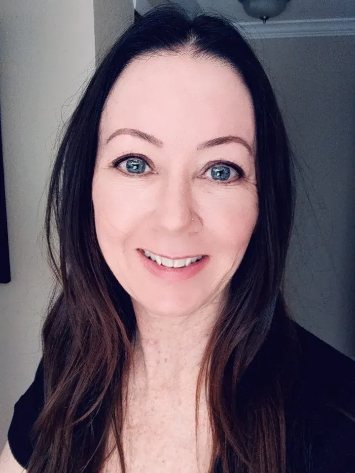 A person with long, dark hair and blue eyes smiling at the camera in a brightly lit indoor setting.