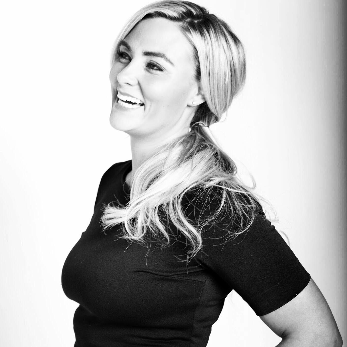 A black-and-white portrait of a smiling person with long hair tied back, wearing a short-sleeved top against a light background.