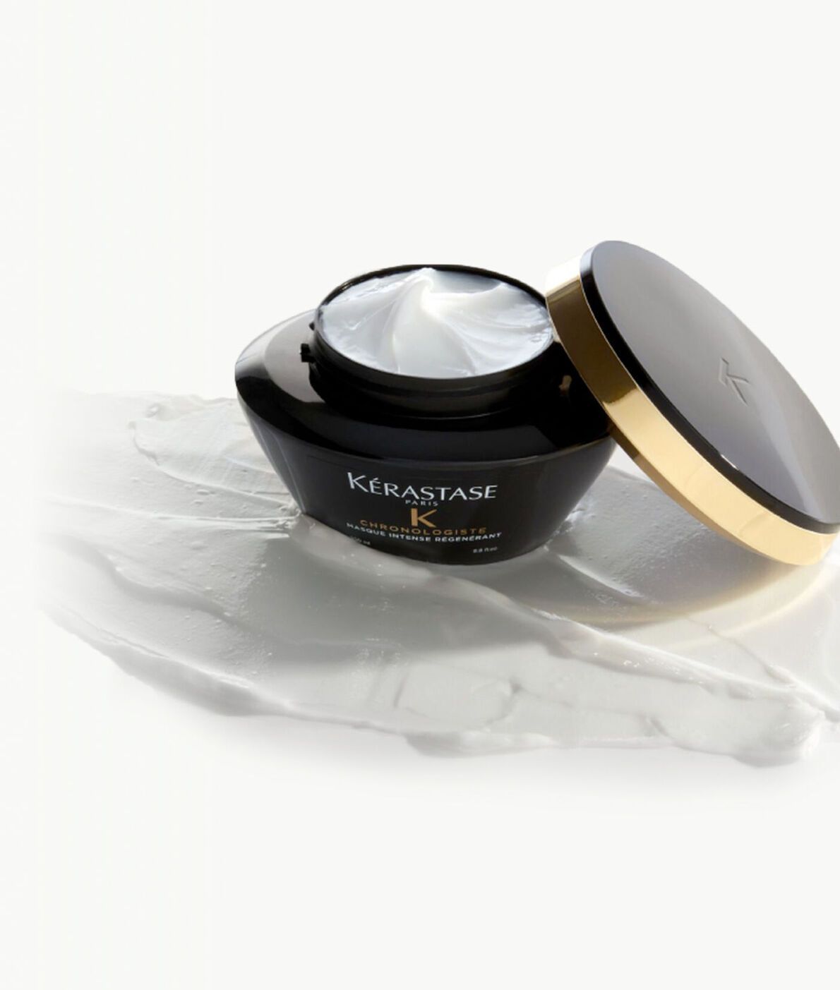 An open black jar of Kérastase Chronologiste hair mask with a gold-rimmed lid resting on a streak of white cream.