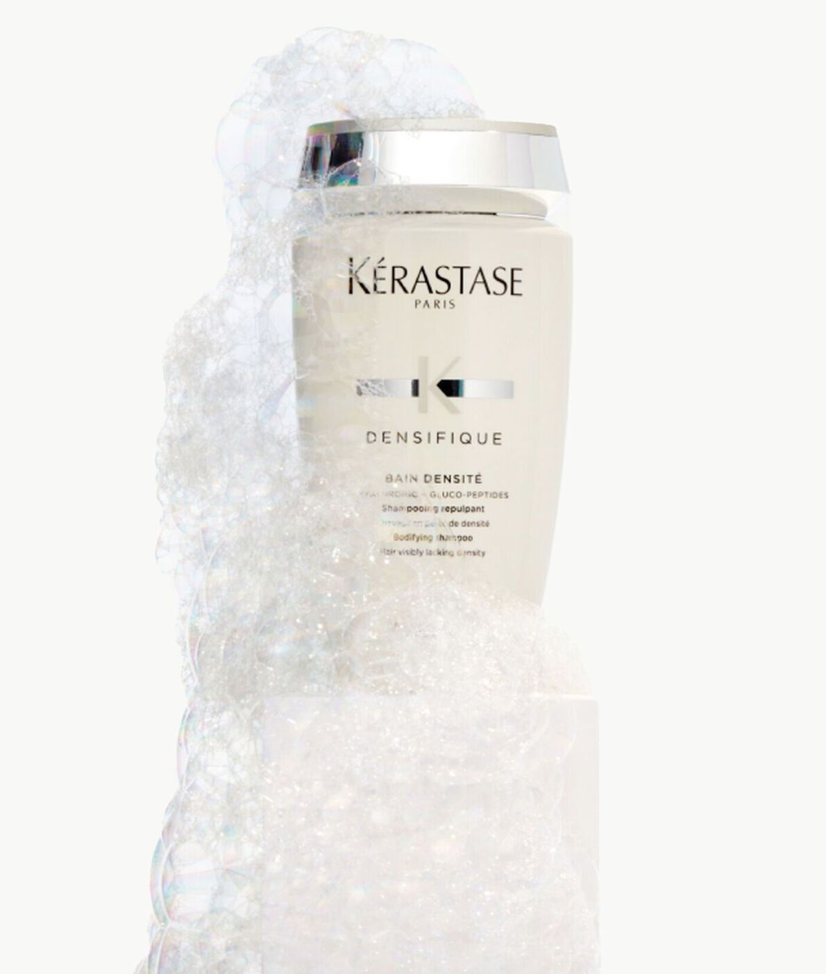 A bottle of Kérastase Densifique shampoo wrapped in bubbly, translucent white foam against a white background.