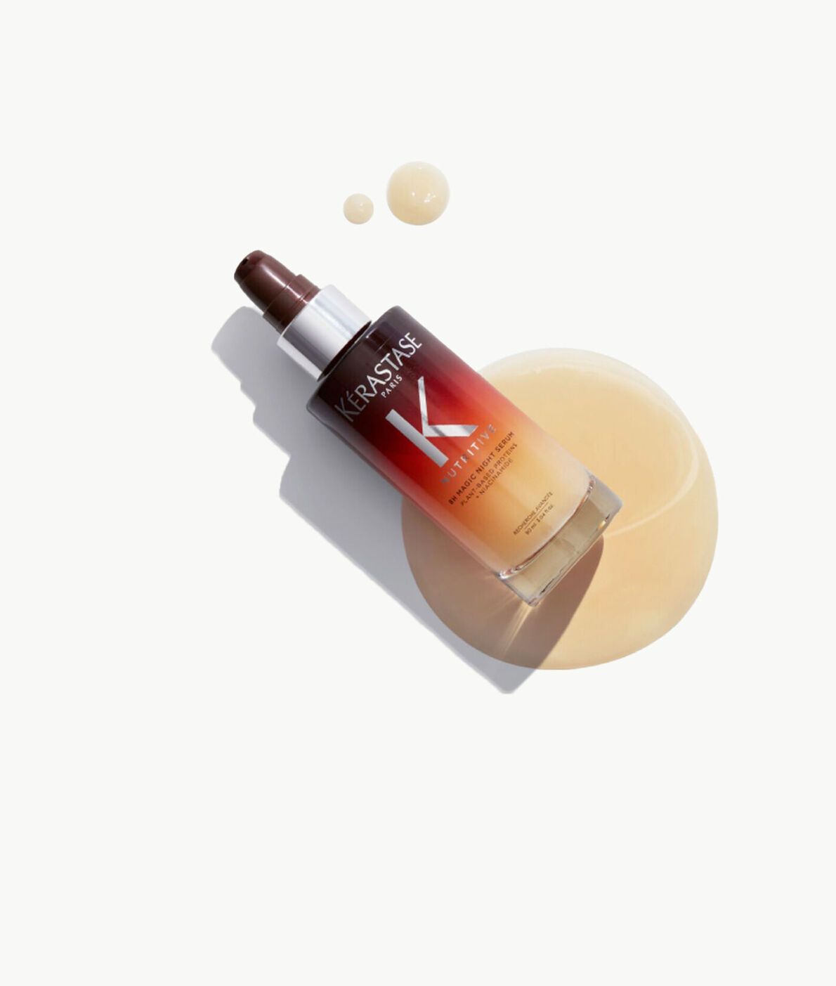 A bottle of Kérastase hair oil with an ombre red and gold design, resting on a pool of spilled oil on a white background.