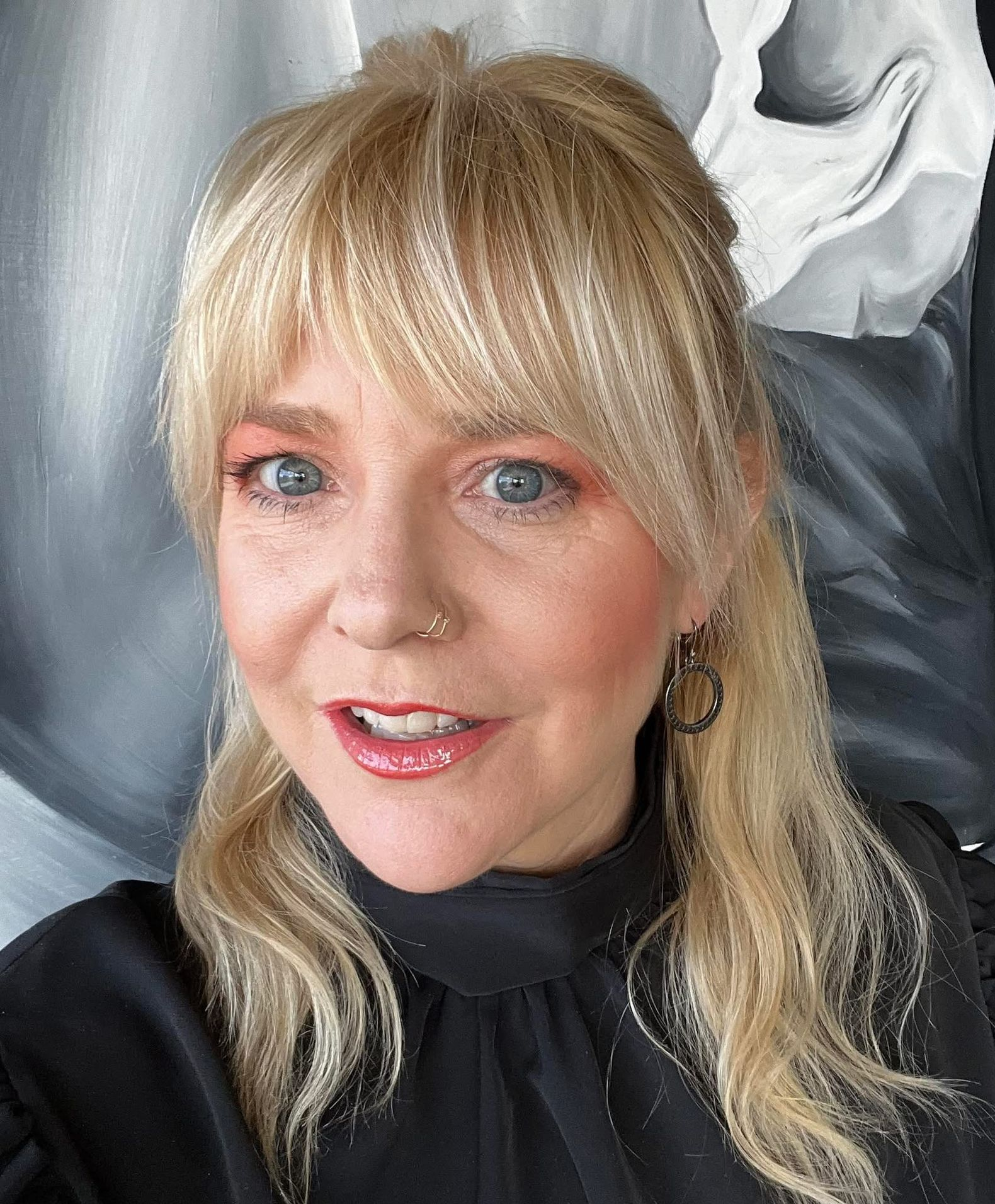 A blonde person with bangs and a nose ring wearing a black top, smiling in front of a grey, abstract background.