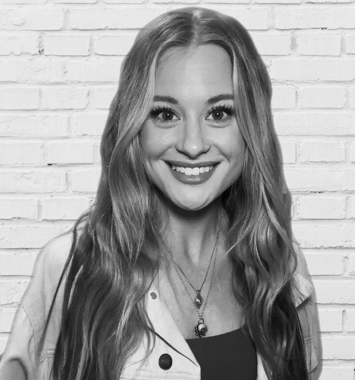 Smiling person with long hair wearing a layered necklace in front of a white brick wall, in black and white.