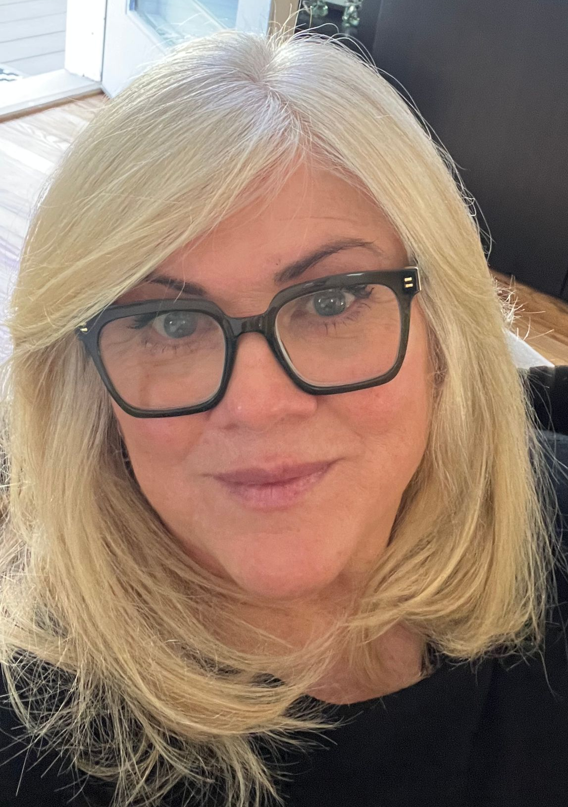 A person with blonde hair and black-rimmed glasses smiling gently at the camera.