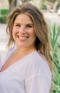 A person with long, light brown hair smiling at the camera, wearing a white V-neck shirt in a garden setting.