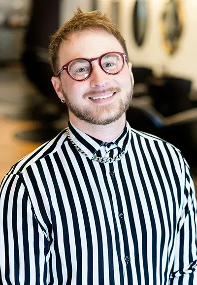 A person wearing reddish-brown glasses and a black-and-white vertical striped button-down shirt, smiling in a salon.