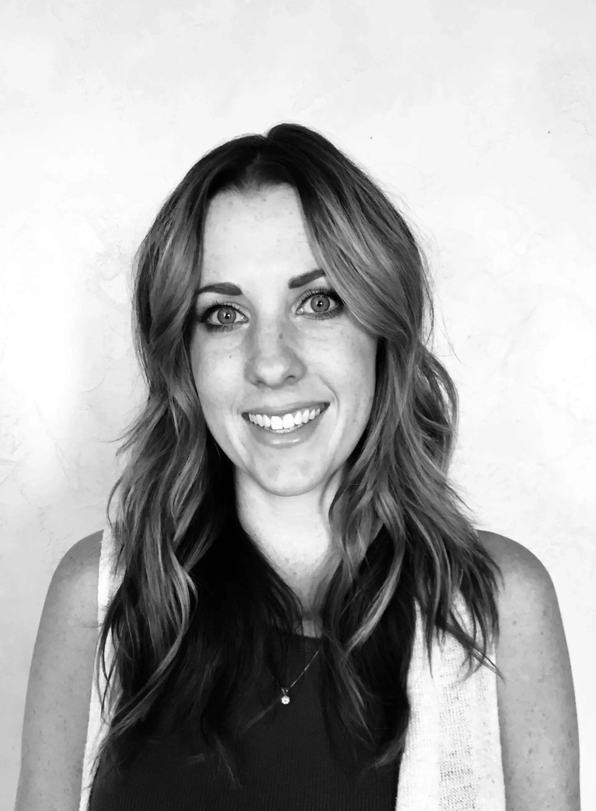 A smiling person with shoulder-length hair and a layered outfit posing against a plain background.