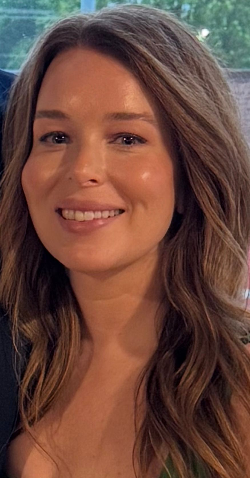 A person with wavy brown hair smiling at the camera, wearing a dark top, with a blurred outdoor background.