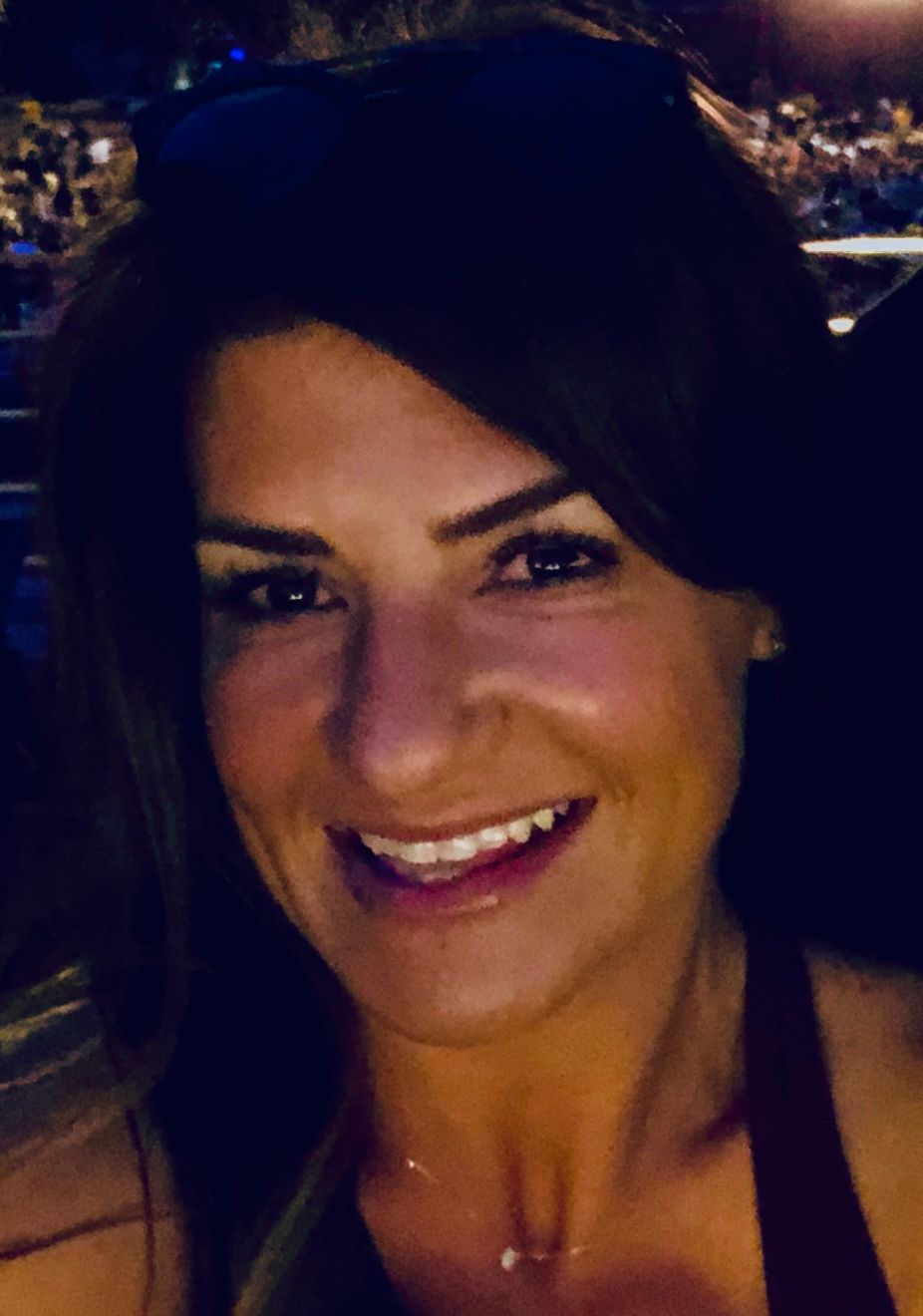 A smiling person with shoulder-length hair and dark sunglasses on their head, captured in a dimly lit, crowded setting.