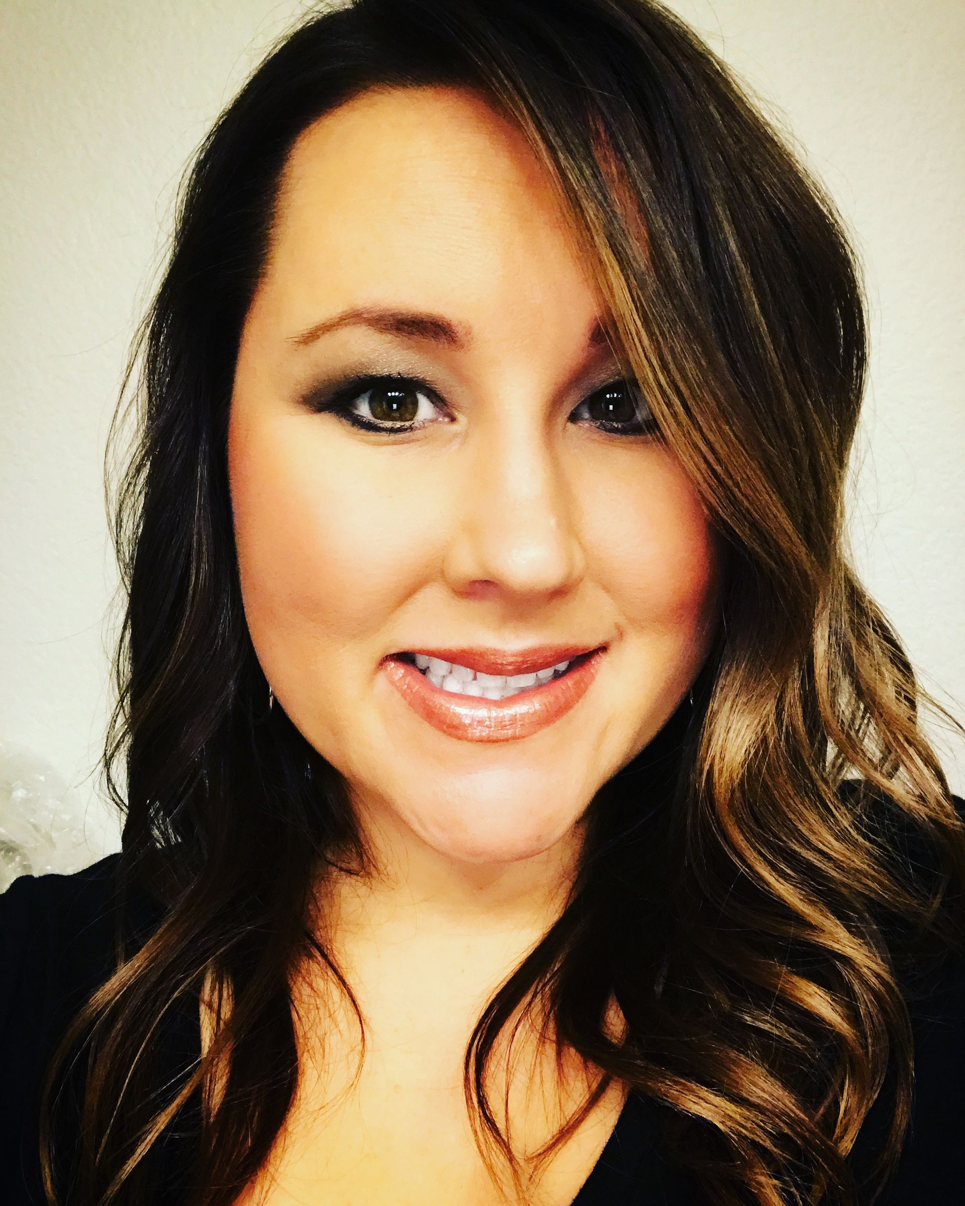 Close-up portrait of a smiling person with long, wavy brown hair, wearing a black shirt against a plain light background.