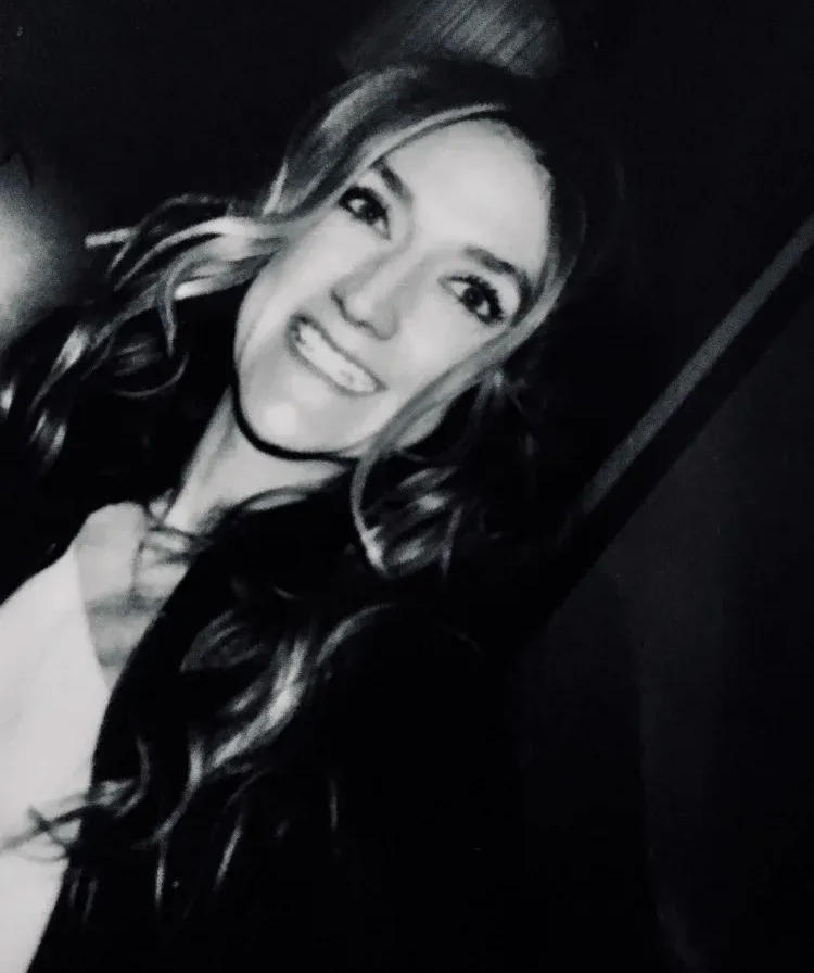 A black-and-white portrait of a person with long wavy hair smiling toward the camera, wearing a light top and dark jacket.