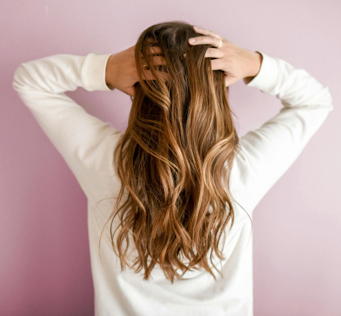 A person from behind with long, wavy brown hair, wearing a white sweater against a pink background, with hands on head.