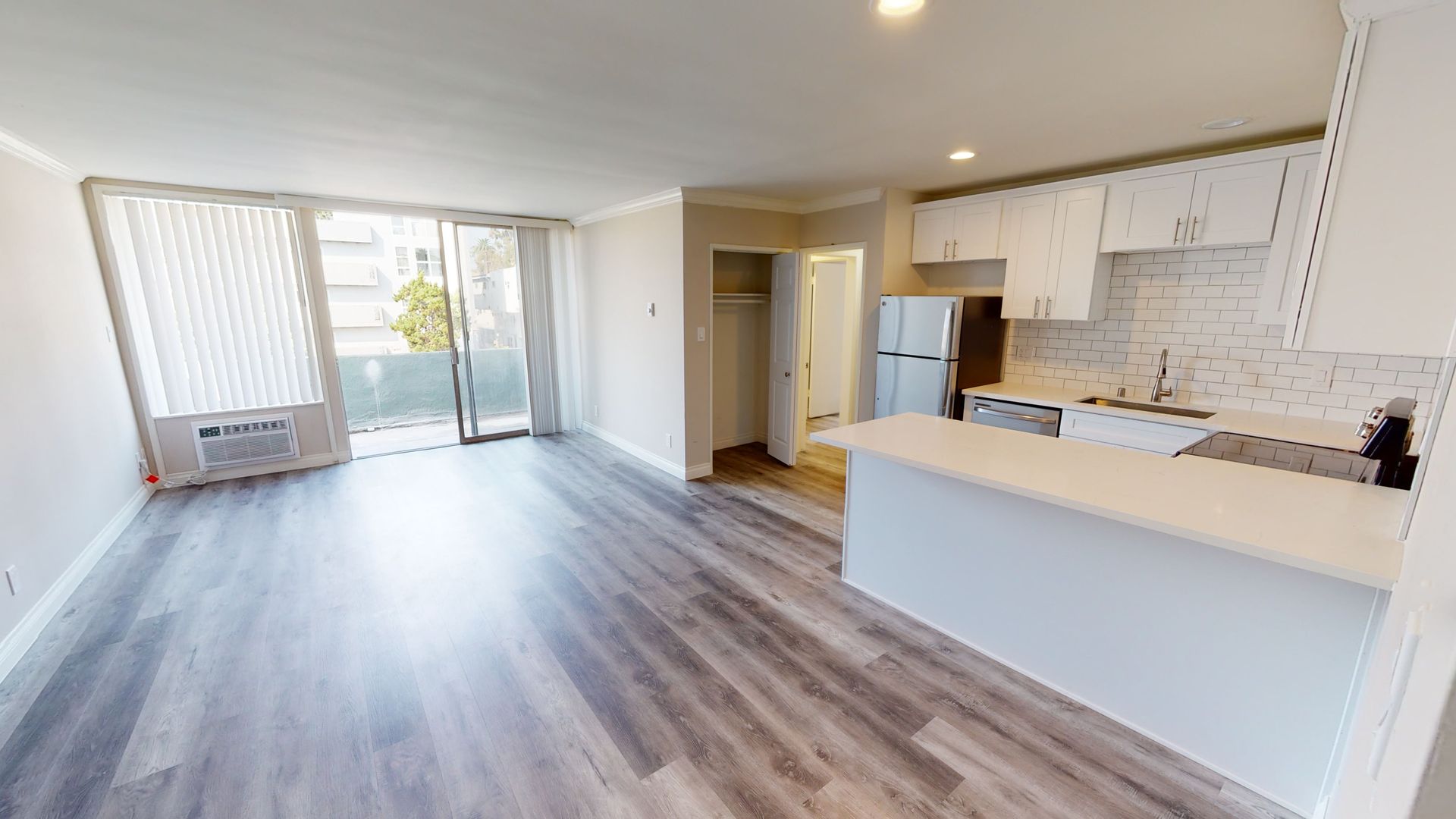 Interior view of a modern apartment with wood floors, white kitchen, and sliding door to a balcony.