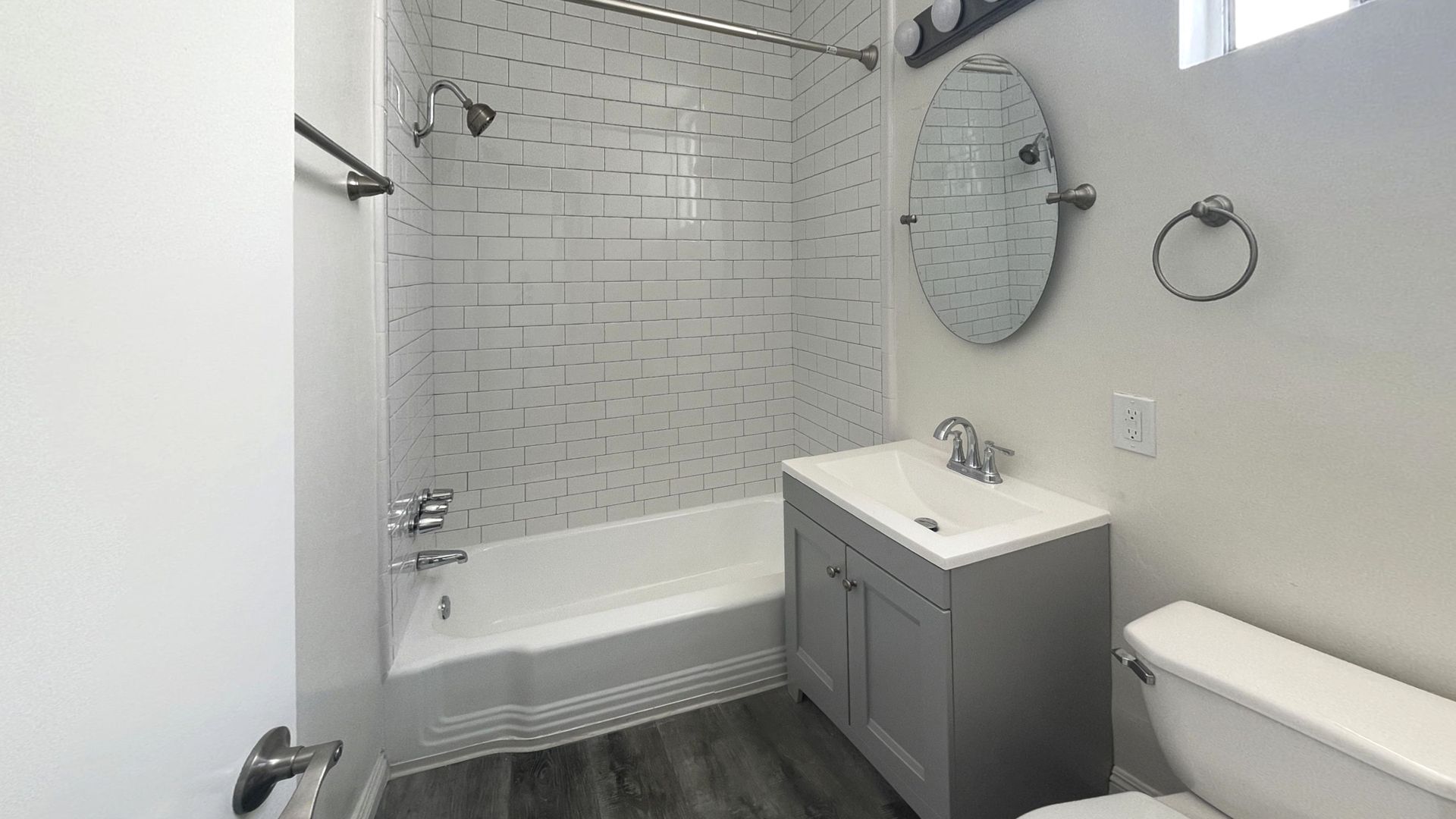 Bathroom with white and gray tiled shower, gray vanity, toilet, and oval mirror.