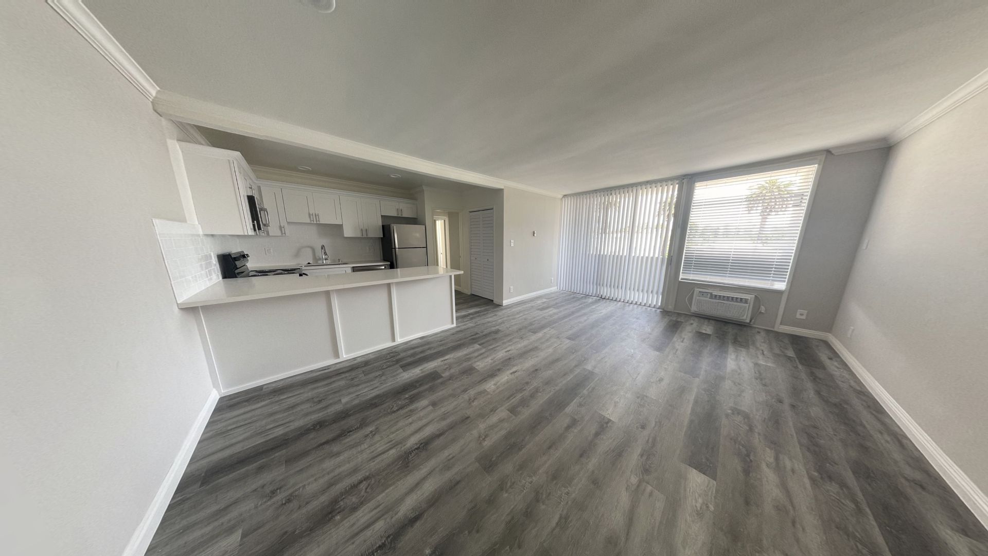 Living room with gray wood floors, white walls, and kitchen area. A window with white curtains and blinds.