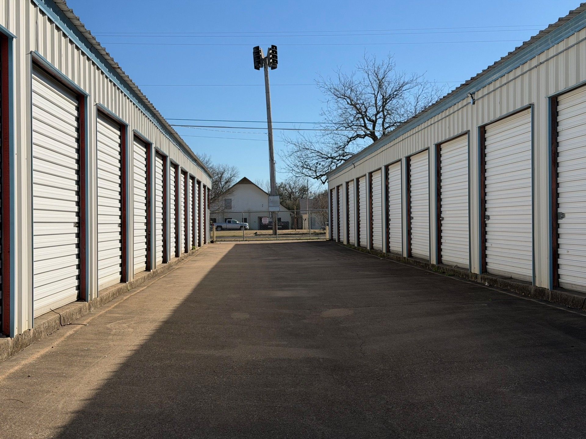 Storage units with white doors line a paved driveway, a utility pole and building in the distance, under a blue sky.