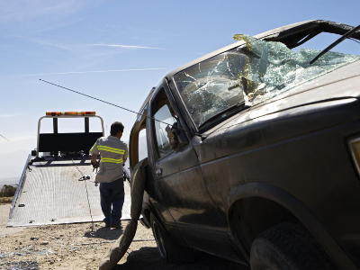 Damaged black truck being towed by a tow truck on a sunny day. A worker in a safety vest is working.