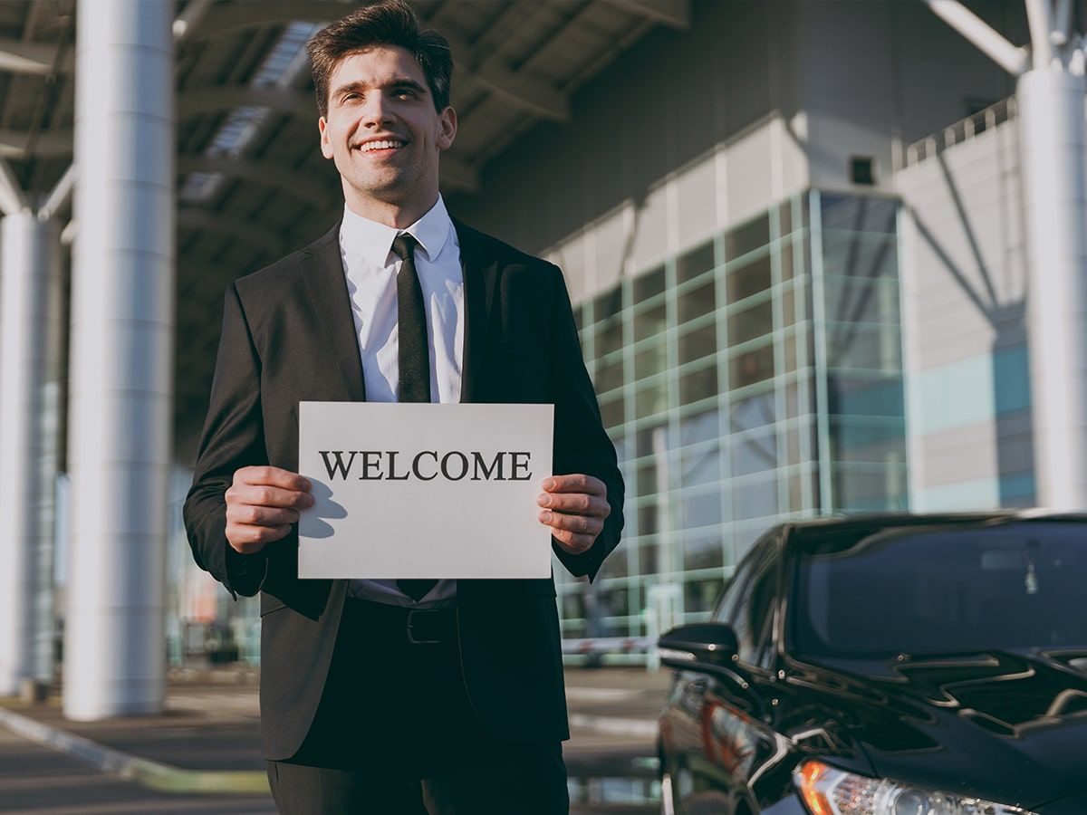 A professional chauffeur stands next to a sleek black luxury sedan outside an airport terminal.