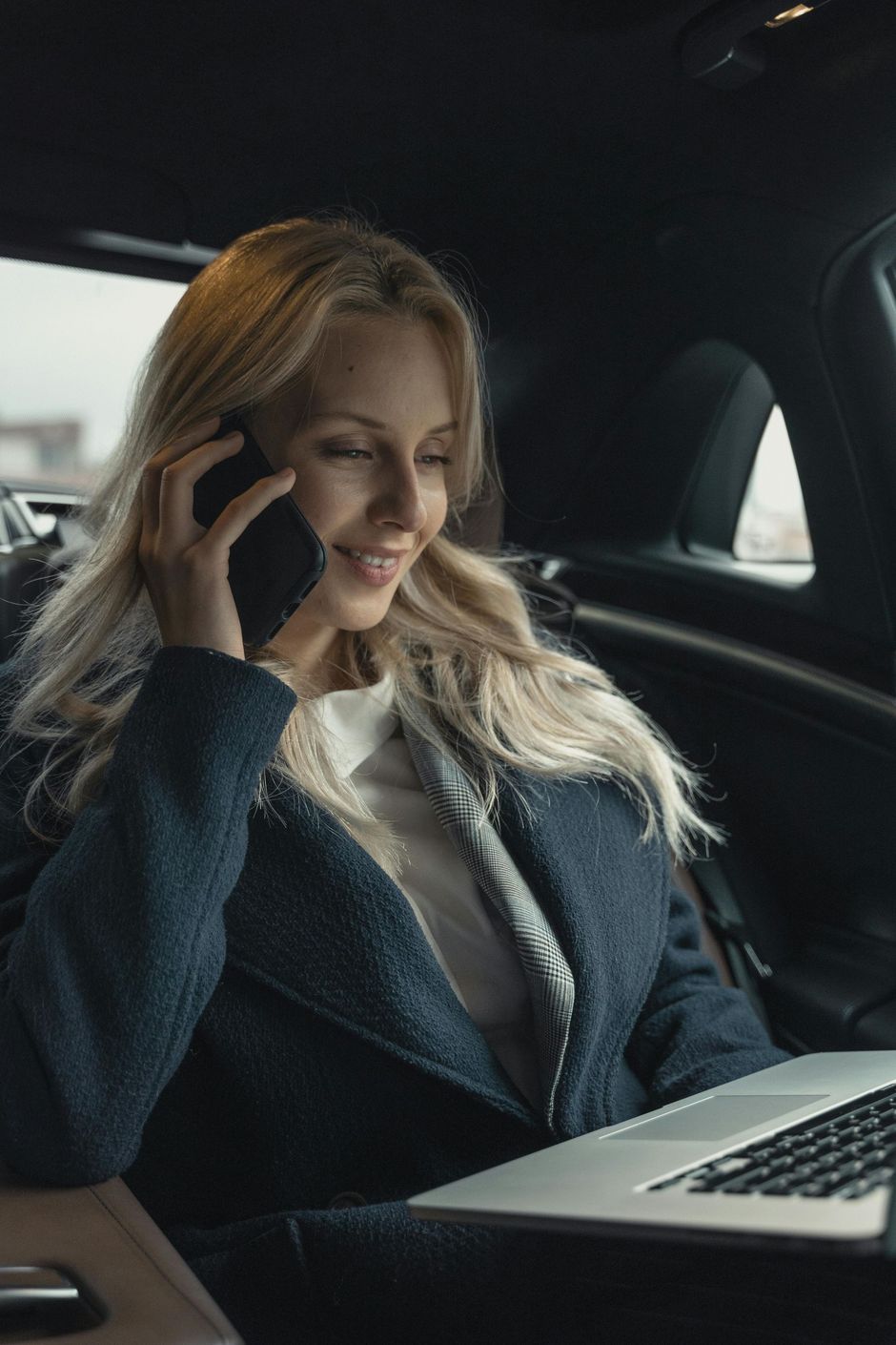 A woman is sitting in the back seat of a car talking on a cell phone while using a laptop.