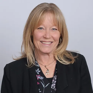 Smiling woman in a black blazer and floral top against a light gray background
