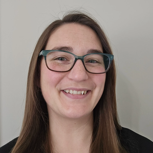 Smiling person with long brown hair and teal glasses against a plain light wall