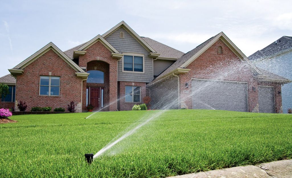 A lawn sprinkler is spraying water on a lush green lawn in front of a large brick house.