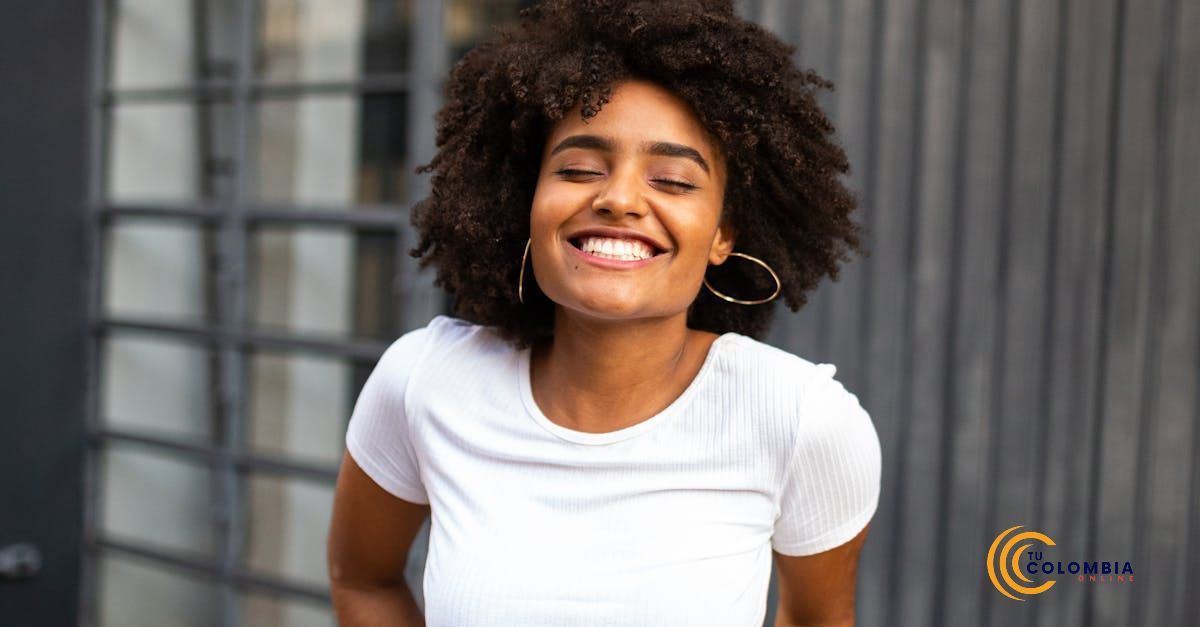 A woman with curly hair is smiling with her eyes closed and wearing a white t-shirt.