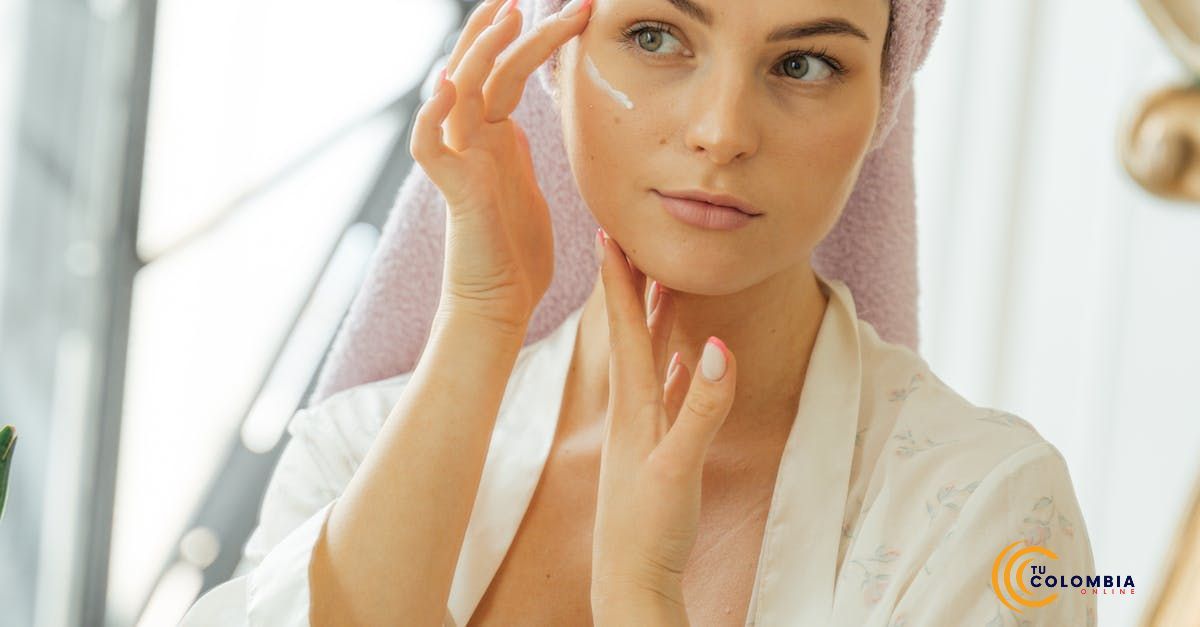 A woman with a towel wrapped around her head is applying lotion to her face.